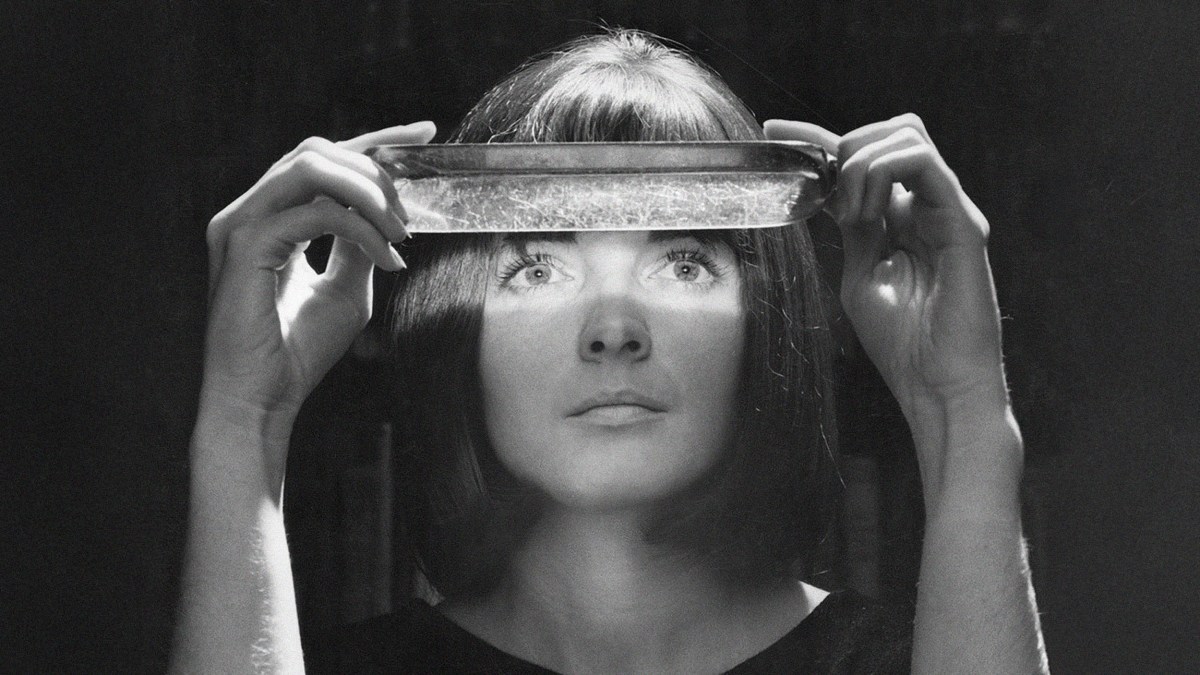 A woman, resembling a paranormal investigator, holds a rectangular glass dish above her eyes, which are illuminated by light shining through the dish in a dark setting.