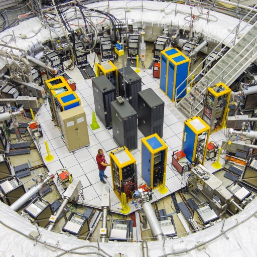 A large circular particle accelerator laboratory with various machines, cables, and equipment; two people are working near the center on experiments related to the muon g-2 anomaly.