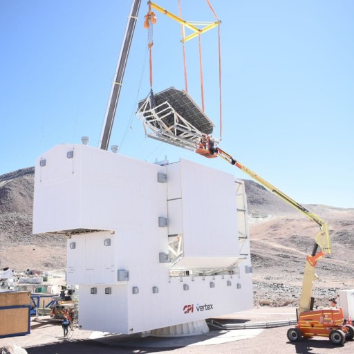 A crane lifts a large metal structure onto a white building at a construction site in a mountainous, arid area under clear blue sky.