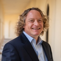 A man with curly blond hair wearing a dark suit and light blue shirt smiles while standing in a sunlit hallway with arched doorways in the background.