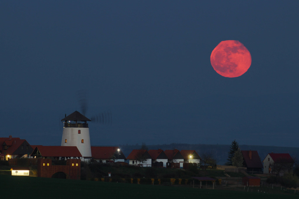 A large red moon rises over a village with a windmill and houses at dusk, some lights glowing in buildings&mdash;an inspiring scene perfect for 5 science lessons about the moon.