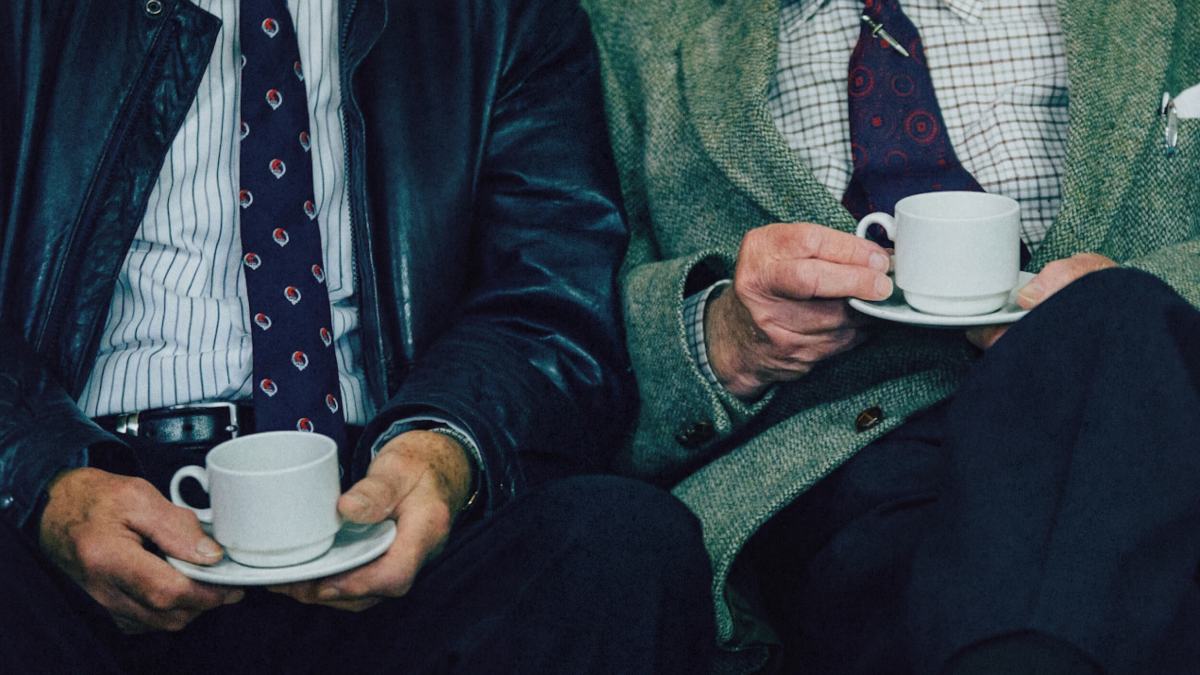 Two men in suits sit side by side, each holding a white cup and saucer, with only their torsos and hands visible—one could easily imagine Aaron Hurst sharing a thoughtful conversation over coffee.