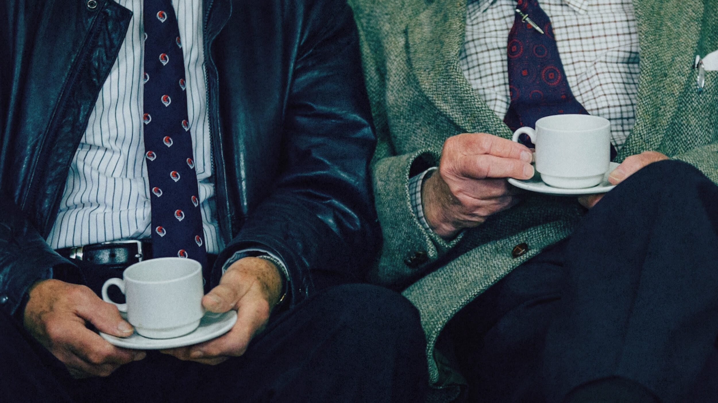 Two men in suits sit side by side, each holding a white cup and saucer, with only their torsos and hands visible—one could easily imagine Aaron Hurst sharing a thoughtful conversation over coffee.