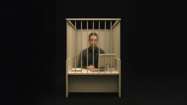 A person sits behind bars at a desk with a computer in a dimly lit setting, suggesting restriction or confinement.