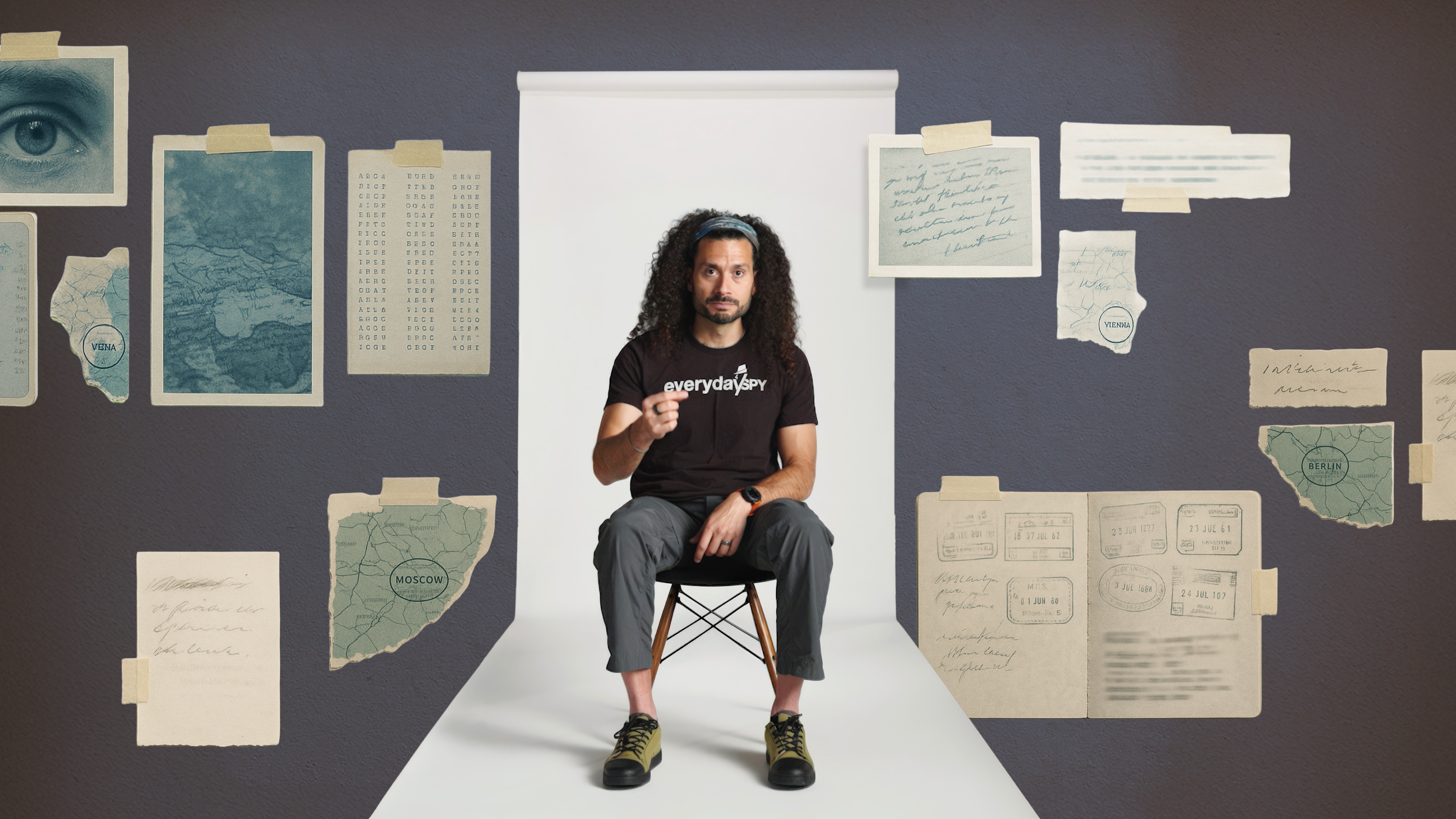 A man with long curly hair sits on a chair in a white hallway, surrounded by illustrated documents, maps, and notes taped to a gray wall.