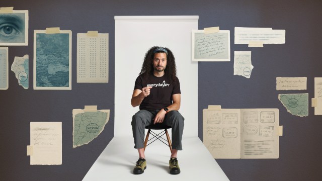 A man with long curly hair sits on a chair in a white hallway, surrounded by illustrated documents, maps, and notes taped to a gray wall.
