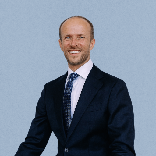 Bogumil Baranowski, dressed in a dark suit, white shirt, and patterned tie, stands and smiles in front of a plain light blue background.