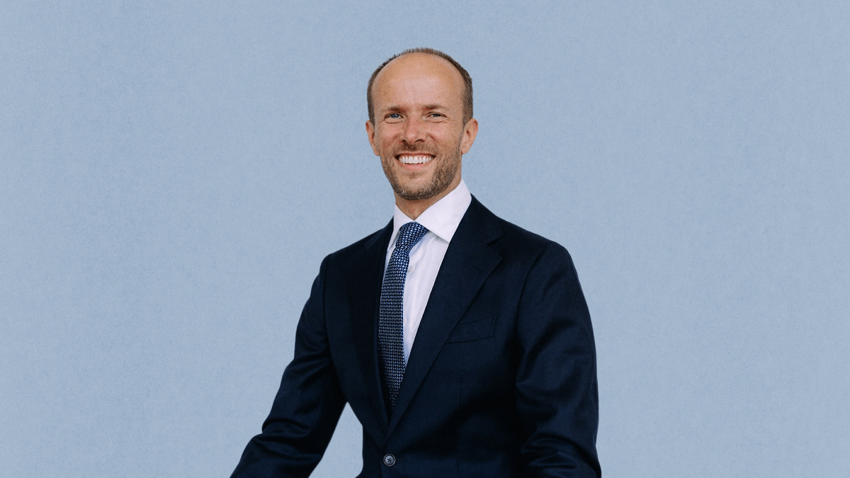 Bogumil Baranowski, dressed in a dark suit, white shirt, and patterned tie, stands and smiles in front of a plain light blue background.