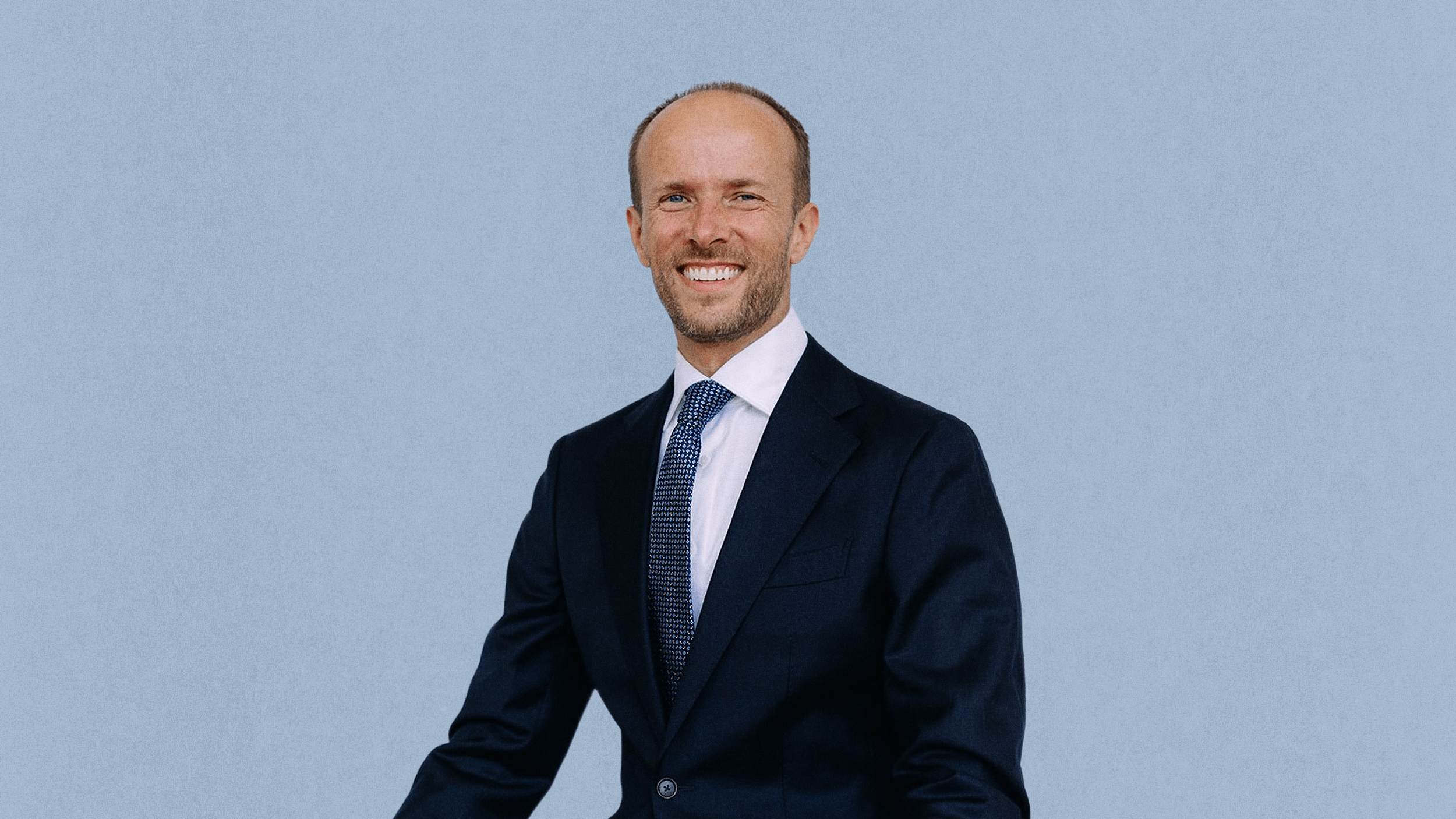 Bogumil Baranowski, dressed in a dark suit, white shirt, and patterned tie, stands and smiles in front of a plain light blue background.