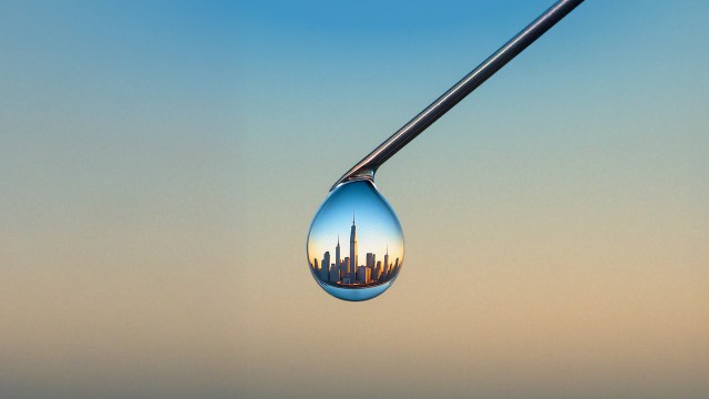 A city skyline is reflected inside a single water droplet hanging from the tip of a metallic object against a gradient sky background.