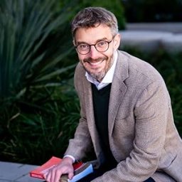 A man with glasses and a beard, wearing a light brown blazer and dark sweater, sits outdoors holding two books, with greenery in the background.
