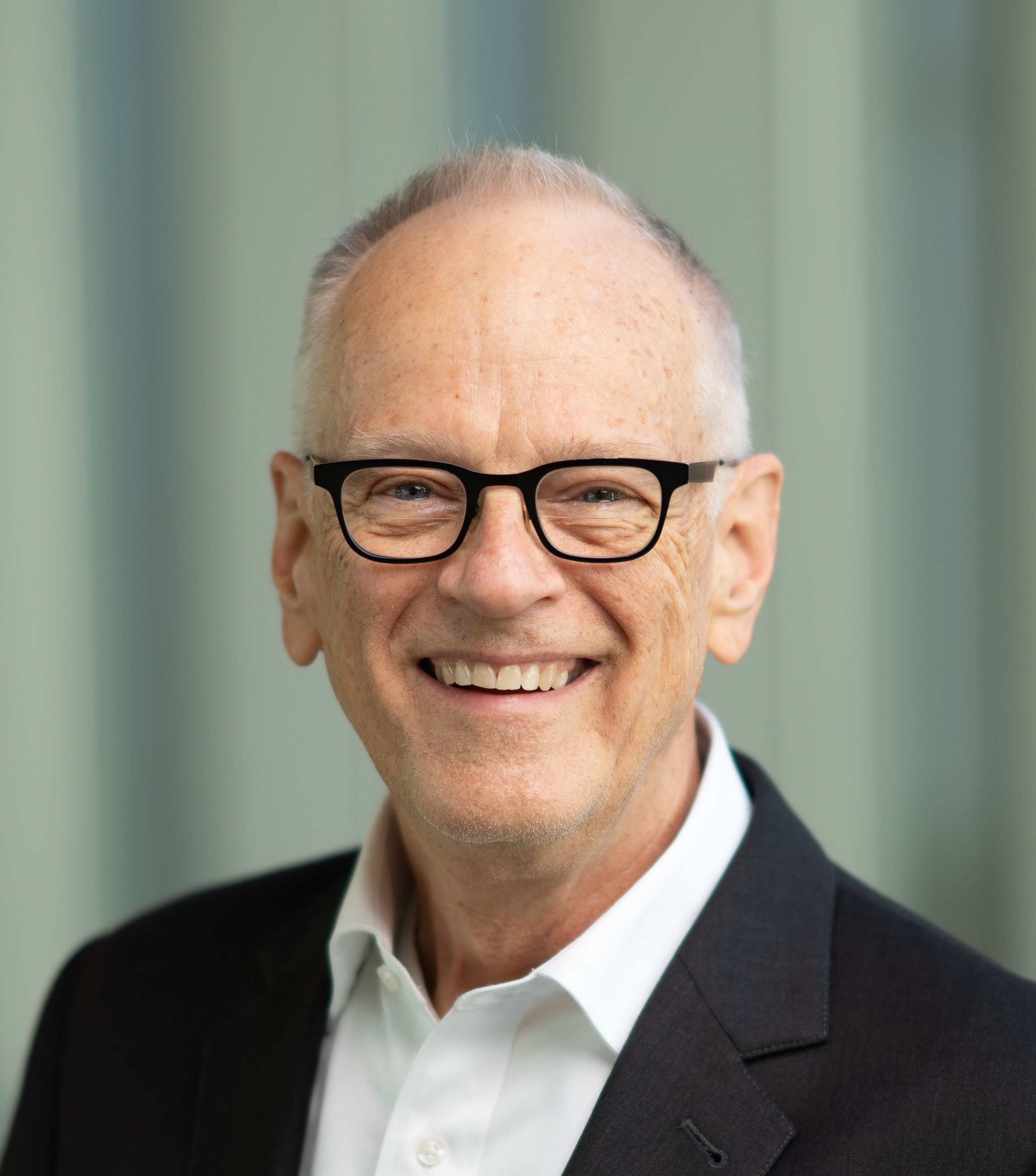Smiling older man with short gray hair and glasses wearing a black suit jacket and white shirt, standing in front of a blurred background.