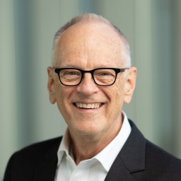 Smiling older man with short gray hair and glasses wearing a black suit jacket and white shirt, standing in front of a blurred background.