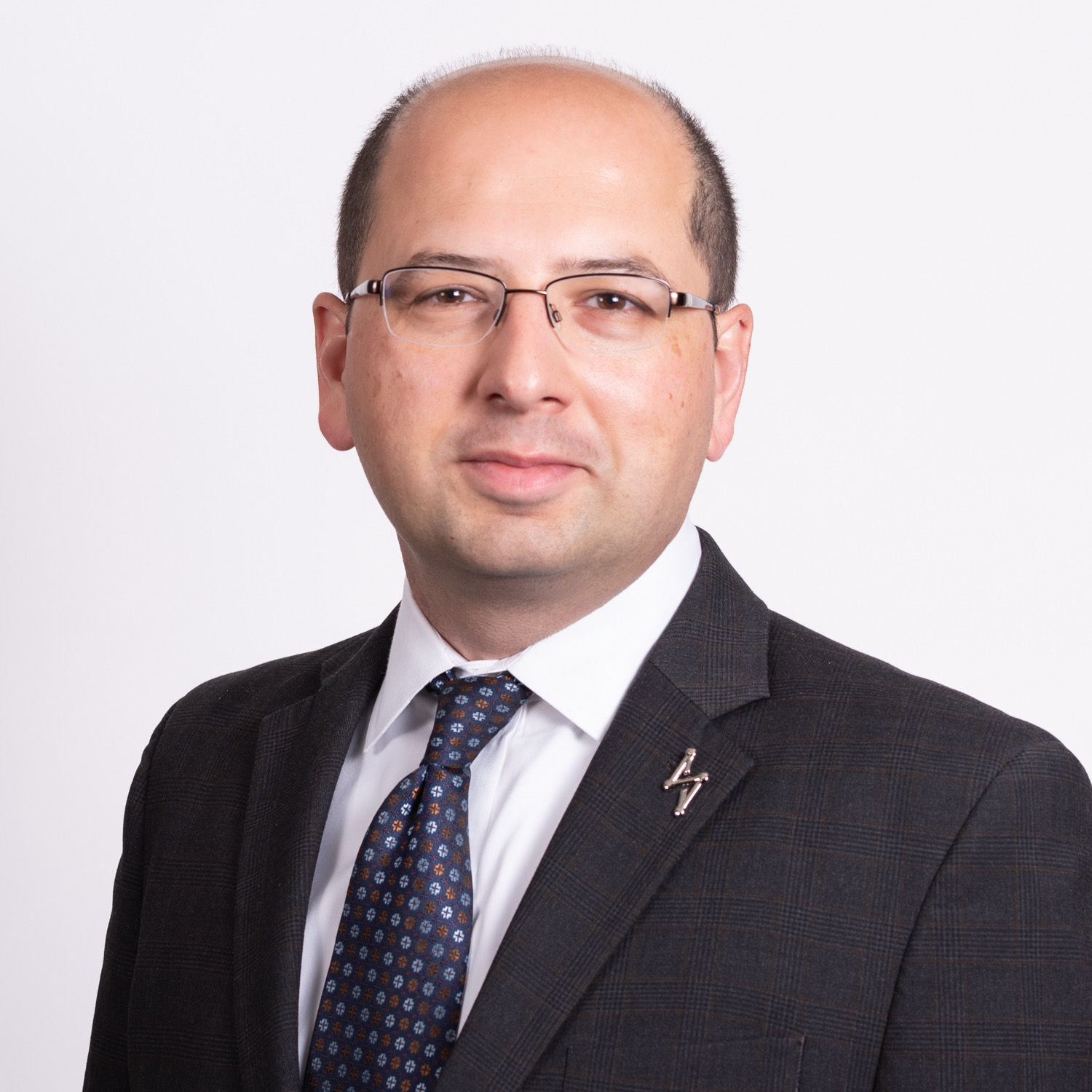A man wearing glasses and a dark suit with a patterned tie poses against a plain white background.