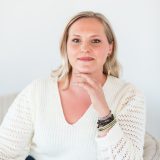 A woman with blonde hair sits in a light-colored chair, wearing a white sweater and jeans, resting her hand on her chin and looking at the camera.