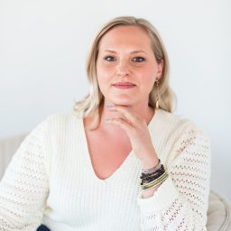 A woman with blonde hair sits in a light-colored chair, wearing a white sweater and jeans, resting her hand on her chin and looking at the camera.