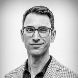 Black and white portrait of a man wearing glasses, a patterned blazer, and a dark shirt, looking at the camera with a neutral expression.