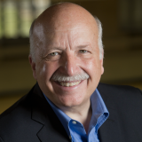 Smiling older man with a bald head, gray mustache, and blue shirt under a dark blazer, posing in front of a blurred indoor background.