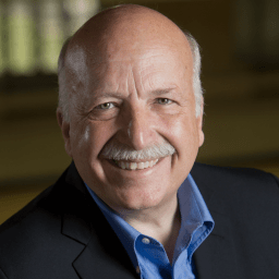 Smiling older man with a bald head, gray mustache, and blue shirt under a dark blazer, posing in front of a blurred indoor background.