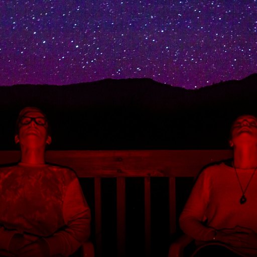 Two people sit on a deck at night, illuminated by red light, looking up at a star-filled sky with mountains silhouetted in the background.