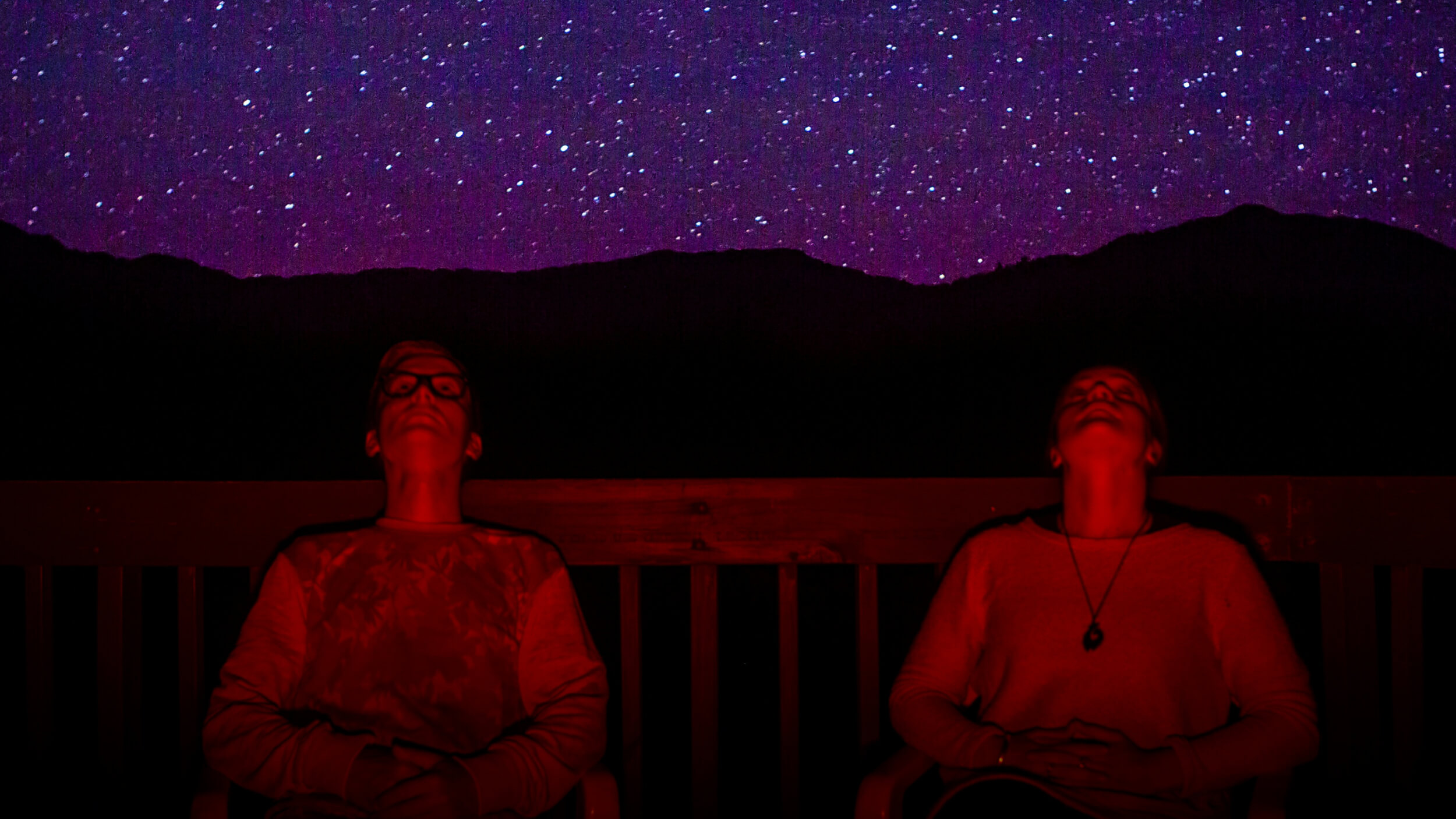 Two people sit on a deck at night, illuminated by red light, looking up at a star-filled sky with mountains silhouetted in the background.