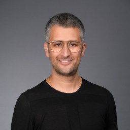 A man with short gray hair and glasses, wearing a black shirt, poses in front of a plain gray background, smiling slightly.