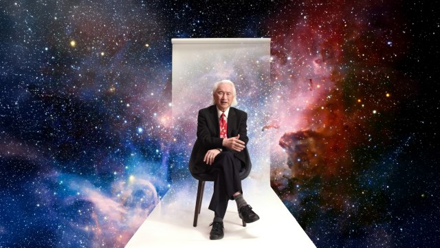 An older man in a suit and red tie sits on a chair against a white backdrop, with a colorful outer space scene in the background.