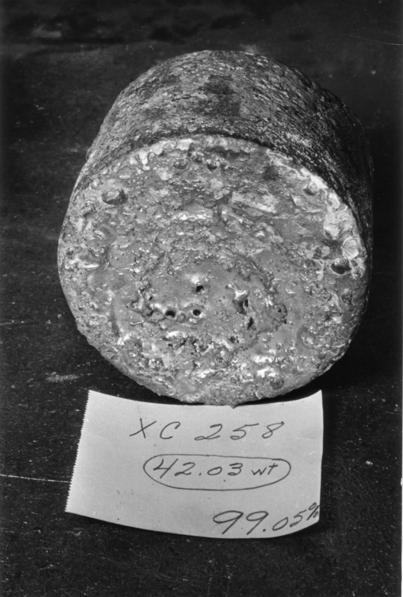 A cylindrical metal ingot sits on a dark surface next to a handwritten note reading &ldquo;XC 258, 42.03 wt, 99.05%.&rdquo;.