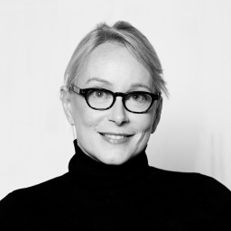 Woman with light hair wearing glasses and a black turtleneck, smiling at the camera against a plain background.