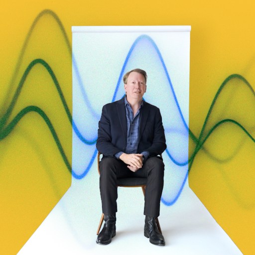 A man in a suit sits on a chair against a yellow background with abstract blue and green wave patterns behind him.