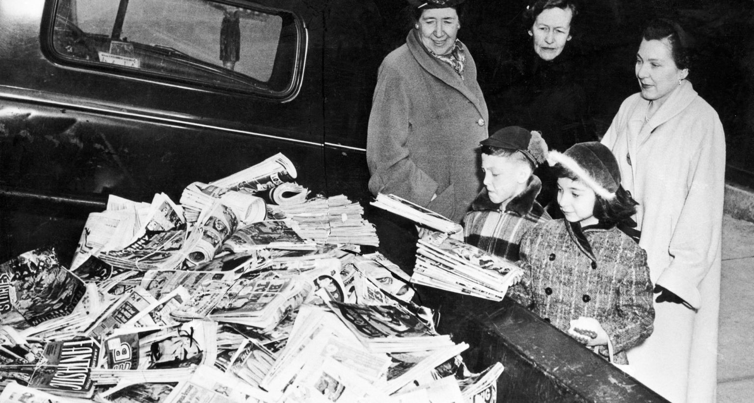 Four adults and two children stand near a large pile of newspapers and magazines outdoors beside a vehicle.