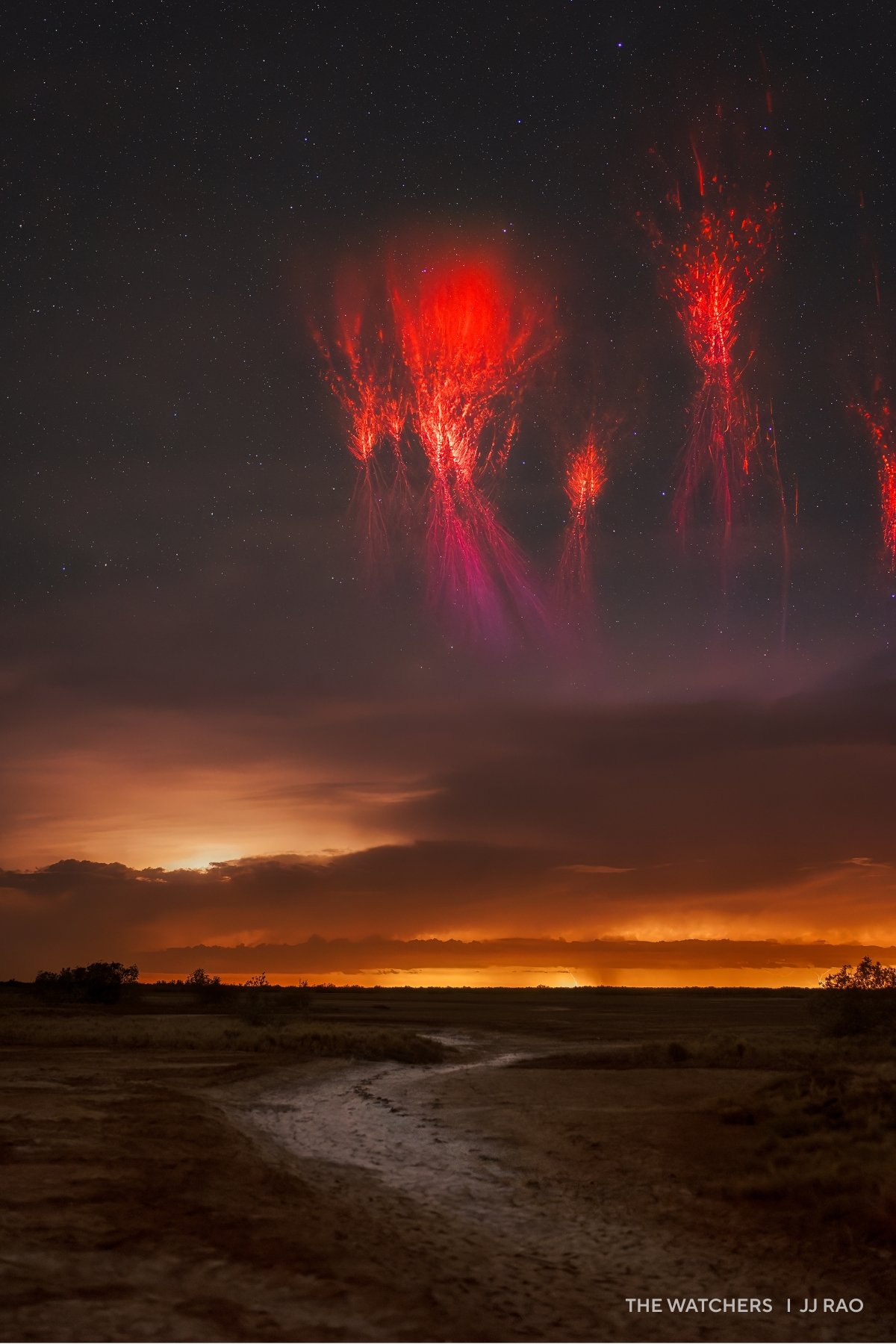 Red sprites illuminate the 2025 night sky above a landscape with a winding stream, silhouetted trees, and a glowing horizon below scattered clouds.