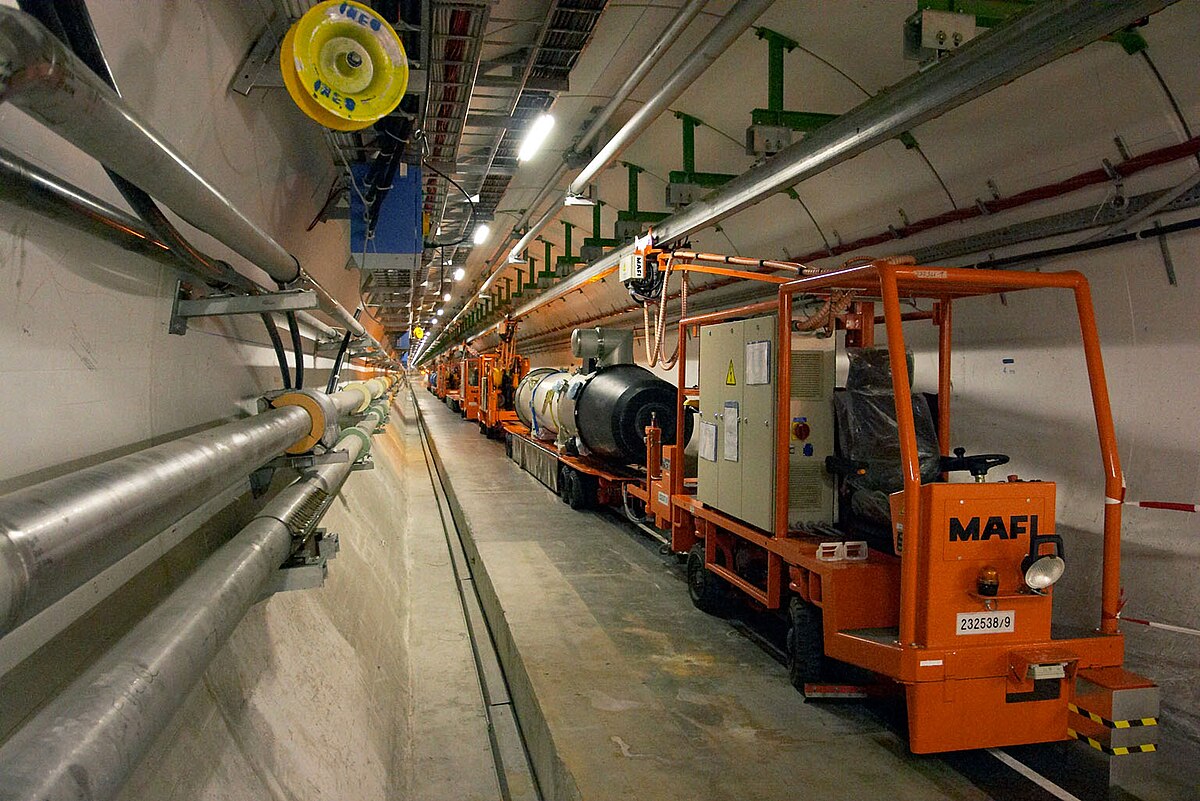 A maintenance vehicle travels along a tunnel next to large pipes and equipment, likely inside a particle physics Higgs factory or similar scientific facility.