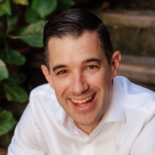 John Candeto, in a white shirt, smiles while sitting outdoors near stone steps and green potted plants.
