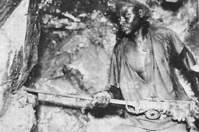 A miner wearing a hat and loose shirt uses a tool to work on a rock face inside an underground mine.