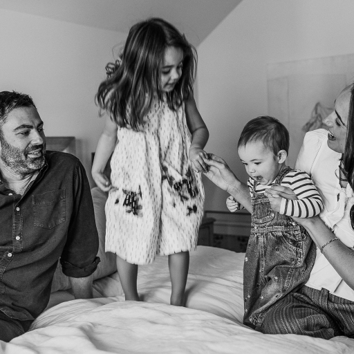 A man and woman sit on a bed with two young children, sharing a quiet moment; the older child stands while the younger, cradled by the woman, embodies the precious gift of consciousness within the family.