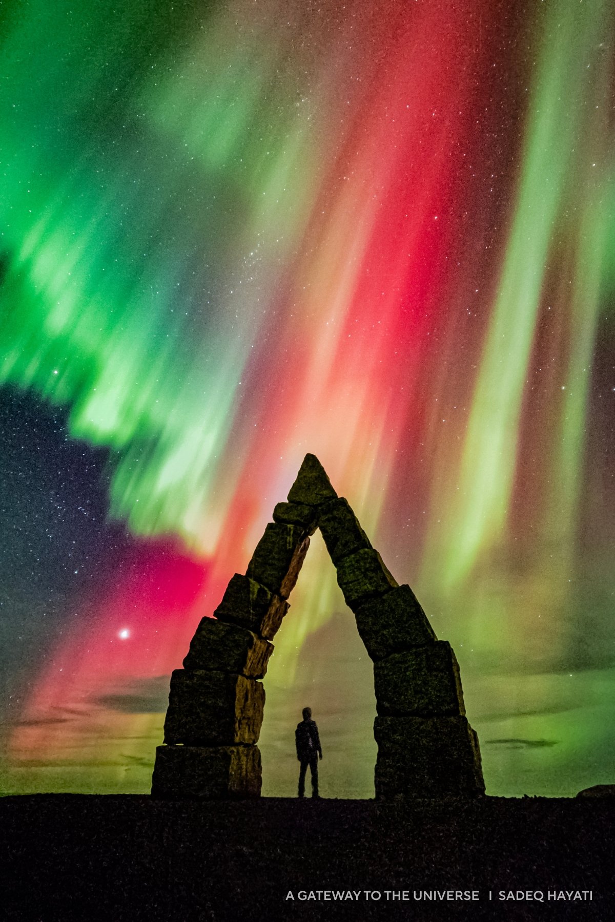 A person stands beneath a stone arch with vivid green and red auroras illuminating the 2025 night sky, creating one of the year's most captivating images.