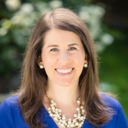 Woman with long brown hair, wearing a blue top, pearl necklace, and earrings, smiling outdoors with greenery in the background.