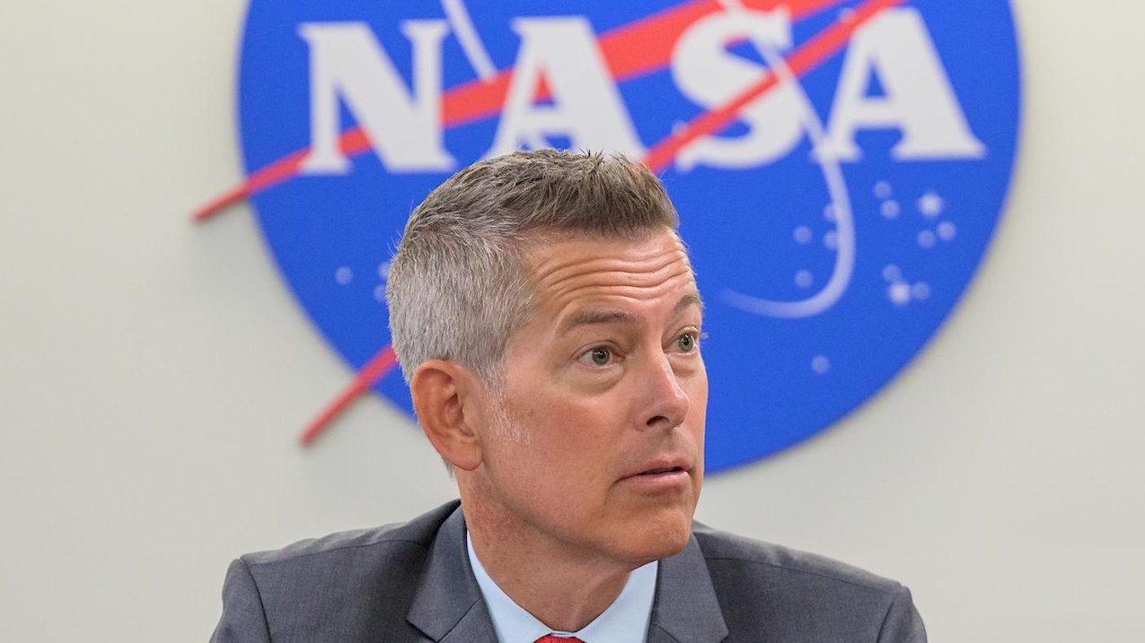 A man in a suit sits in front of a large NASA logo sign, symbolizing the determination to defy charter limits and push the boundaries of space exploration.