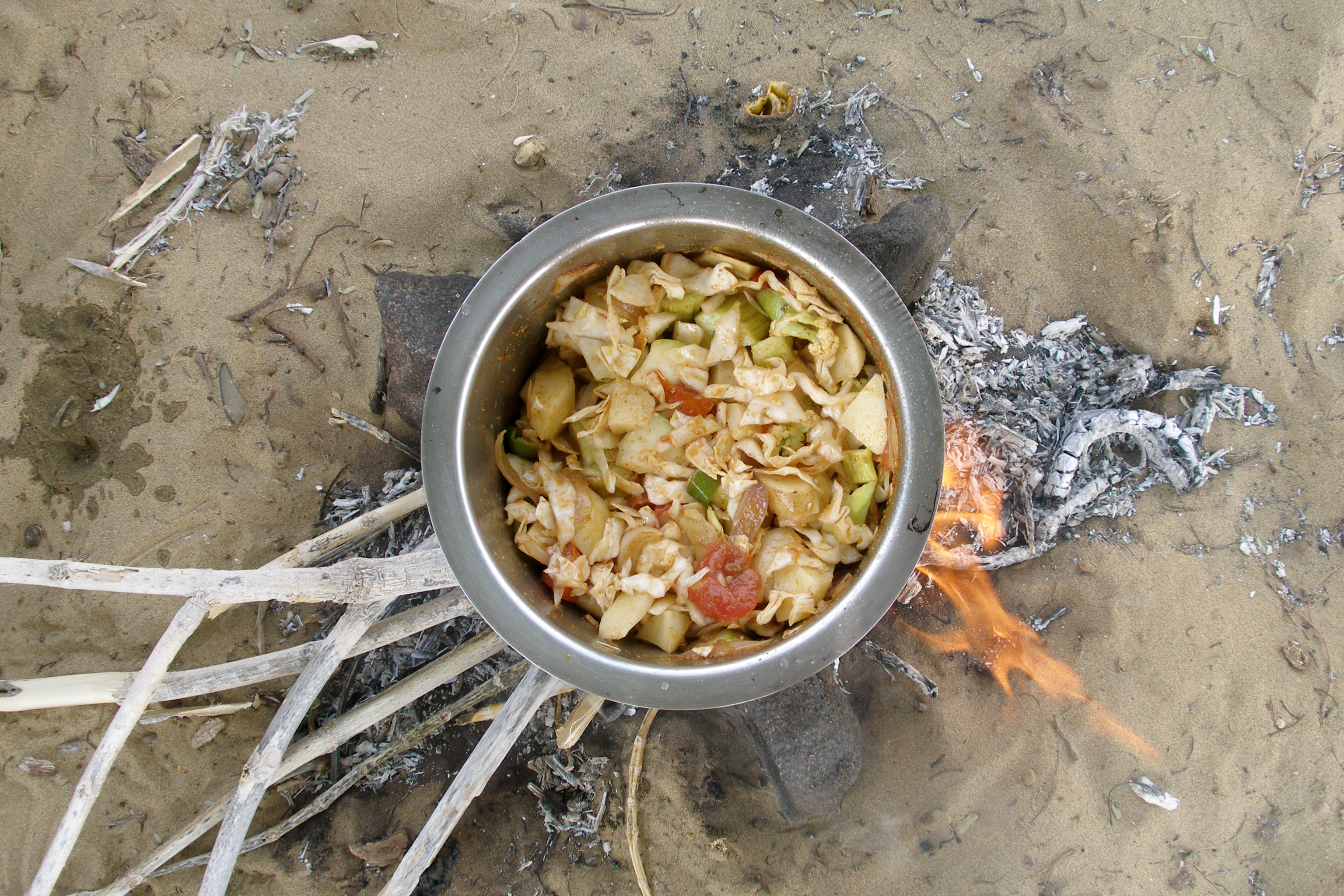A metal pot with vegetables, including cabbage and tomatoes, is cooking over an open fire on sandy ground with scattered ashes and wood.