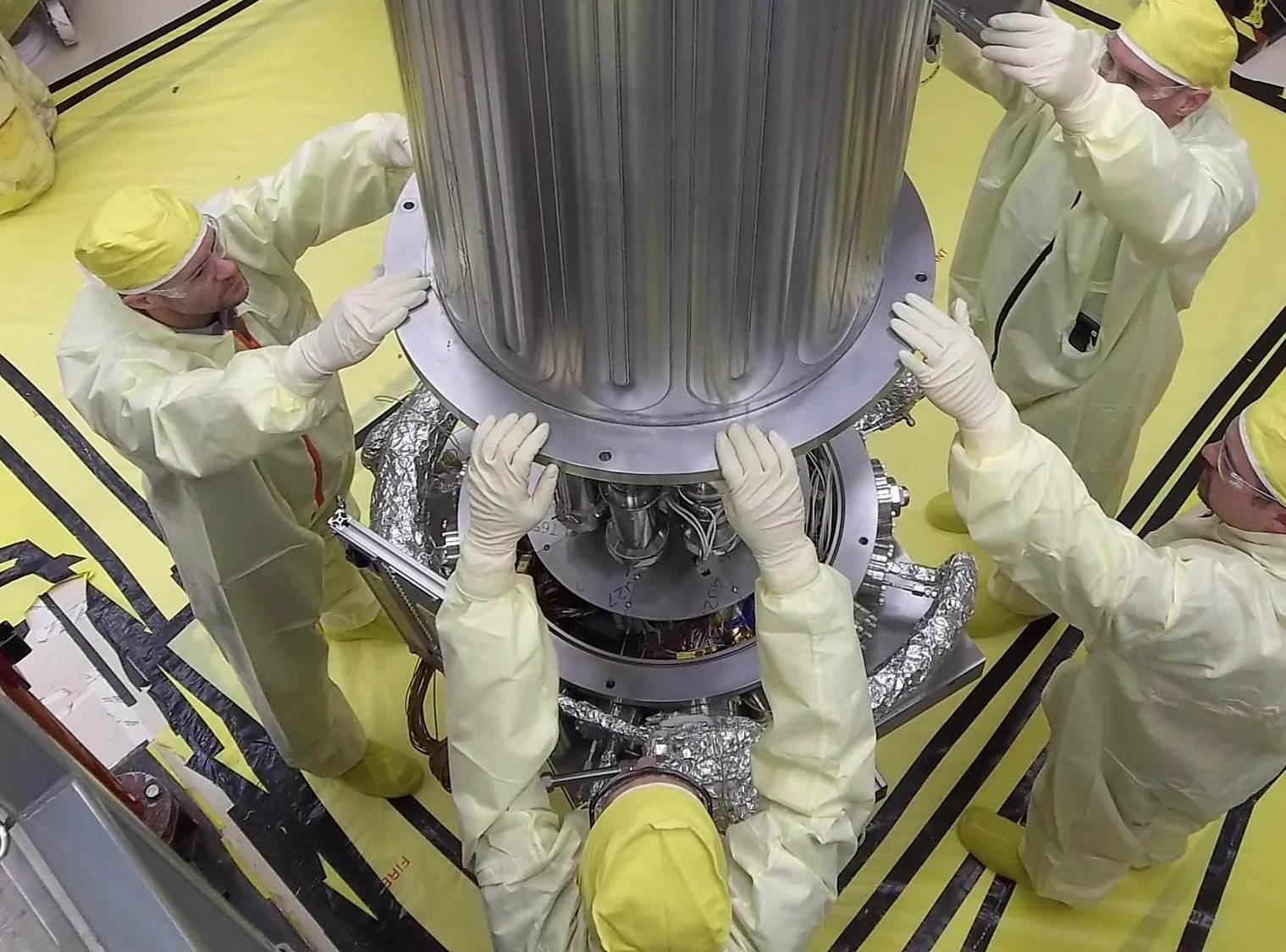 Four technicians in yellow protective suits work together to position a large metallic cylindrical component, resembling part of a nuclear reactor, on a yellow-covered floor during a NASA project.