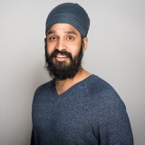 Man with a dark beard and mustache wearing a blue turban and blue sweater, standing against a plain light gray background, looking at the camera and smiling.