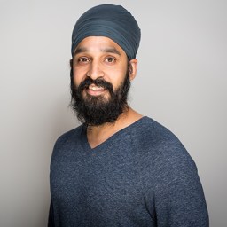 Man with a dark beard and mustache wearing a blue turban and blue sweater, standing against a plain light gray background, looking at the camera and smiling.