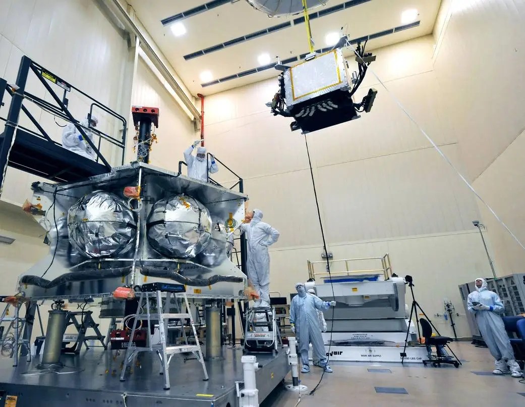 Technicians in protective suits work in a clean room, guiding a satellite component&mdash;part of NASA's Juno mission&mdash;being lowered by a crane onto a larger spacecraft structure.