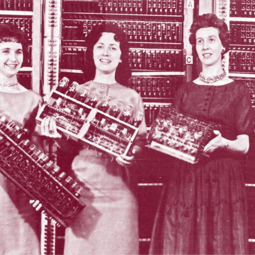 Four women stand in front of a vintage computer, each holding different components of its hardware, with the computer's panel visible in the background—an image reminiscent of historical innovation studied by Angus Fletcher.