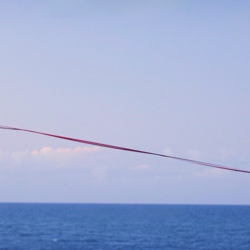 A person walks barefoot on a slackline stretched high above the ocean, with one arm raised for balance and cliffs visible at each end.