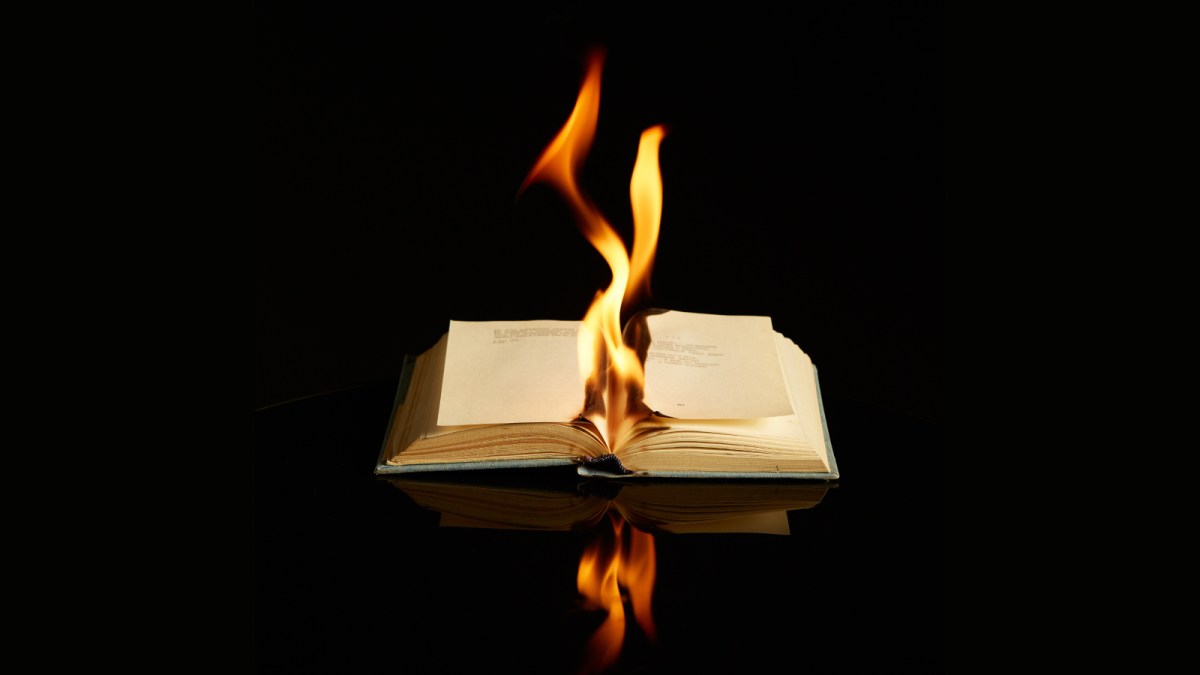 An open book, symbolizing dangerous books, burns with flames rising from its pages against a black background, its fiery reflection shimmering on a glossy surface.