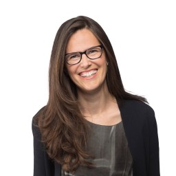 Woman with long brown hair and glasses smiles at the camera, wearing a dark jacket over a patterned top, against a white background.