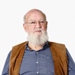 An older man with glasses, a white beard, and a brown vest over a blue shirt, sitting against a plain light background.