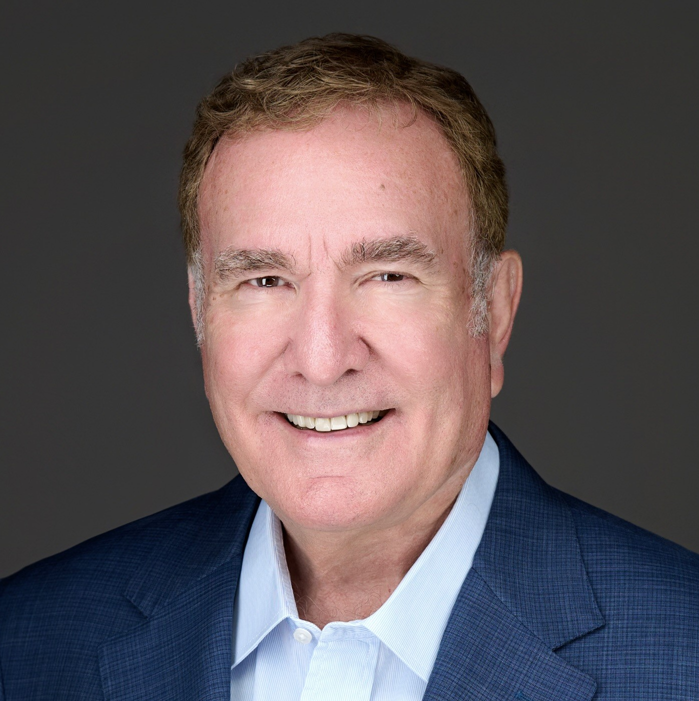 Smiling older man with short light brown hair wearing a blue blazer and light blue collared shirt against a plain dark background.